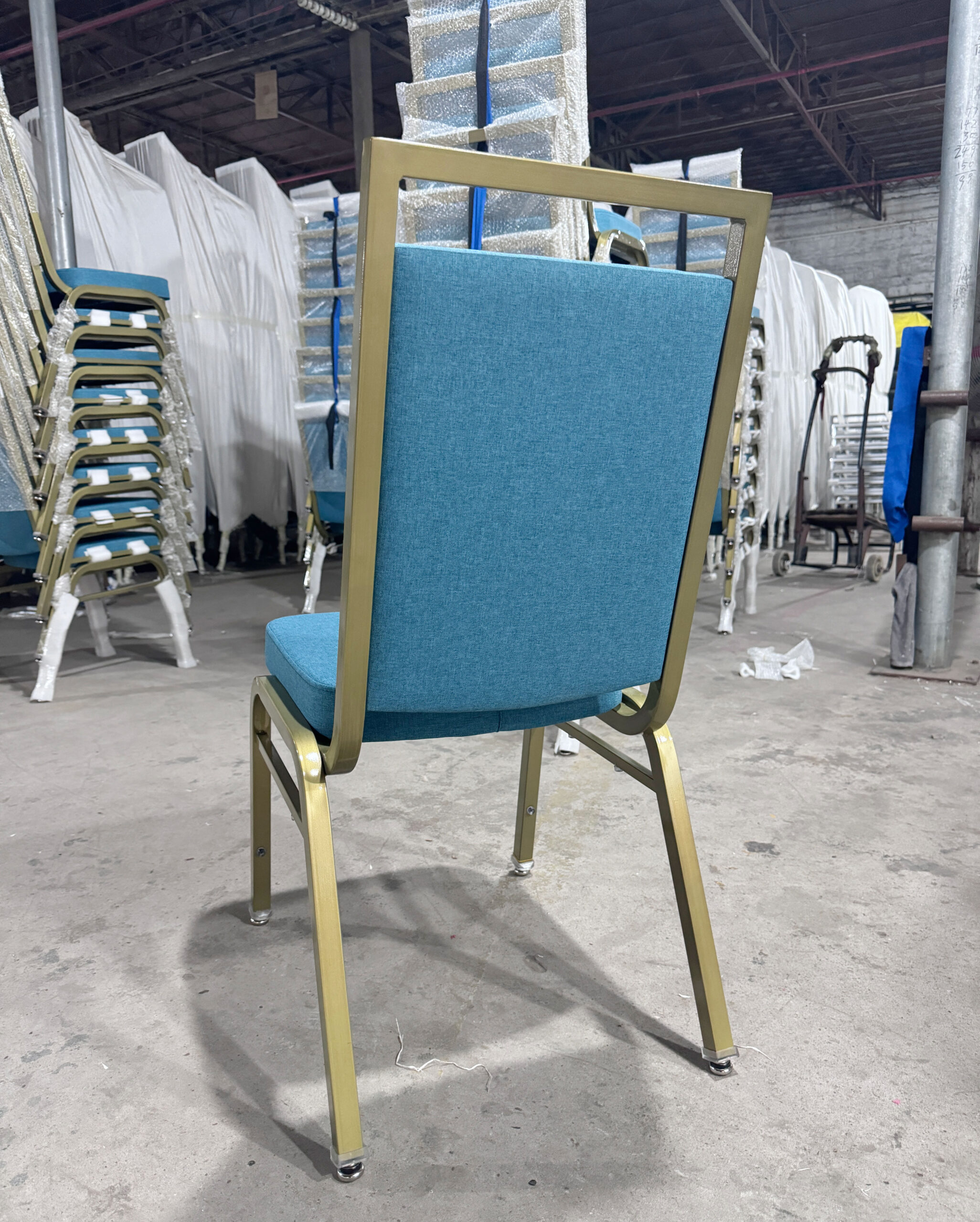 Angled rear view of Valenza banquet chair with blue upholstered backrest and gold powder-coated steel frame, in factory setting