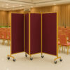 Burgundy banquet hall partition with golden frame and wheels, folding acoustic room divider placed in a hotel banquet dining hall with chairs and curtains.
