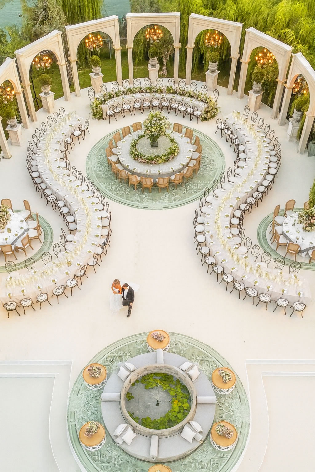 Aerial view of outdoor wedding reception featuring white serpentine banquet tables arranged in flowing curves around a central floral round table