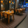Outdoor dining area featuring wooden tables paired with black plastic rattan chairs in a commercial restaurant setting.