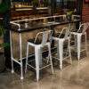 Industrial pipe bar table with white metal bar stools in a modern café interior, featuring a black wood tabletop and commercial-grade steel pipe frame.